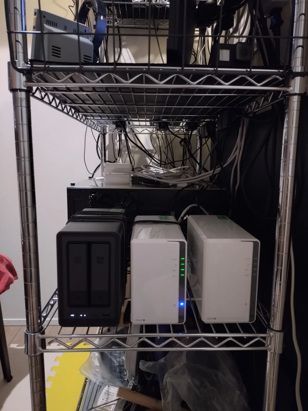 筆者宅の納戸2帖のメタルラックに設置された3台のNAS:Synology DS223j(現役・LED稼働中)・UGREEN NASync DXP2800(現役・黒)・Synology DS216j(前世代機・保管中)と、それぞれに搭載されたNAS用HDDを長期運用中
