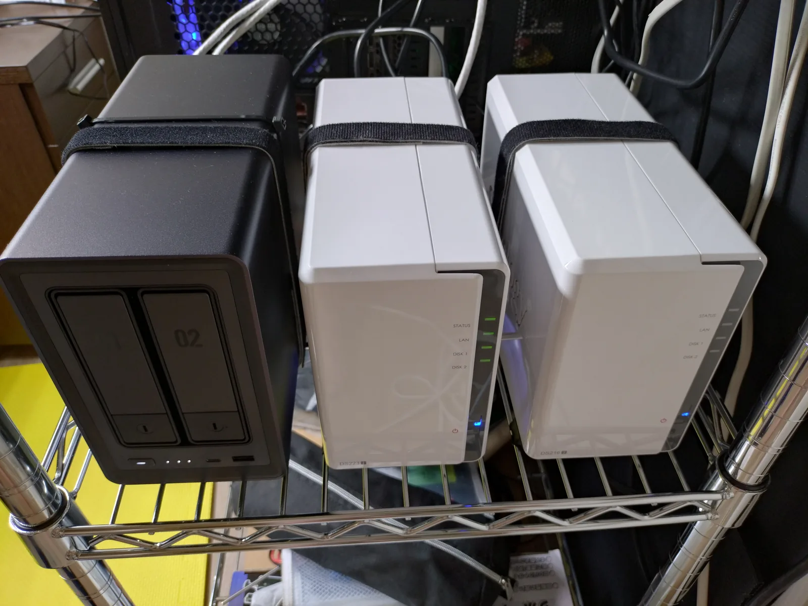 メタルラックに並ぶ2ベイNAS 3台。左からUGREEN NASync DXP2800、Synology DS223j、Synology DS216j。右端のDS216jは電源LED（青）が点灯し通電している
