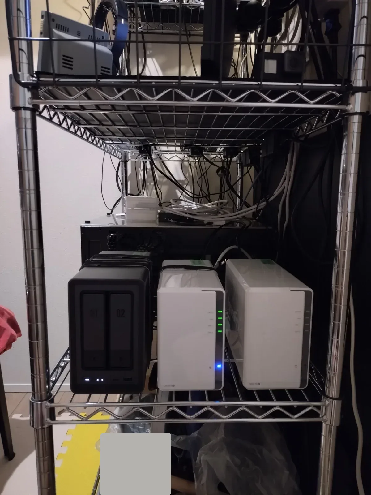 納戸のメタルラックに並ぶ2ベイNAS 3台。左がUGREEN NASync DXP2800（黒）、中央がSynology DS223j（LED点灯）、右端がSynology DS216j（LED全消灯）