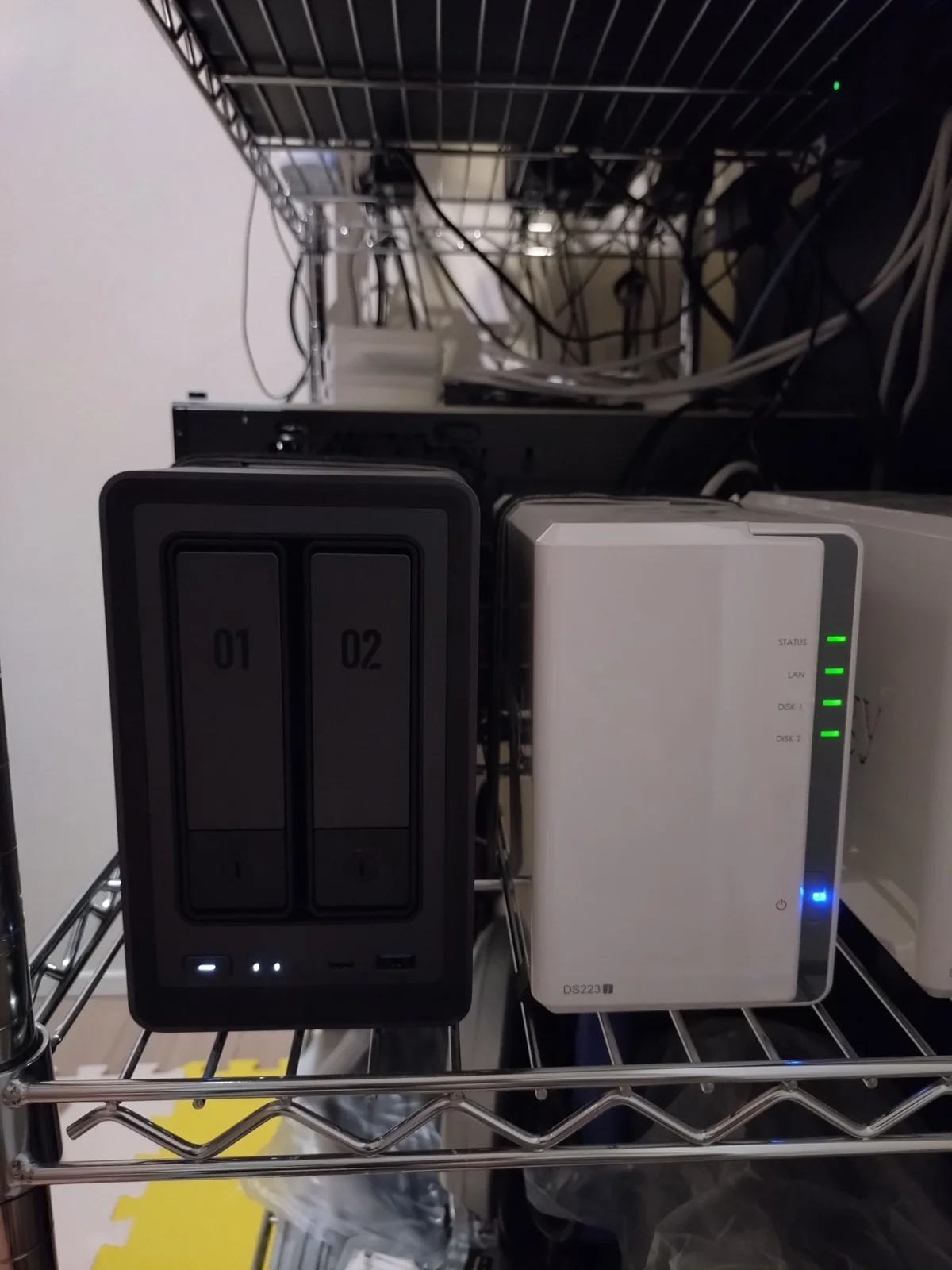 Synology DS223j（右・白・STATUS/LAN/DISK1/DISK2の4個LEDが緑点灯・電源LEDも青点灯）を筆者宅のメタルラックで24時間稼働中の実機写真。左には比較用のUGREEN DXP2800も並んでいる